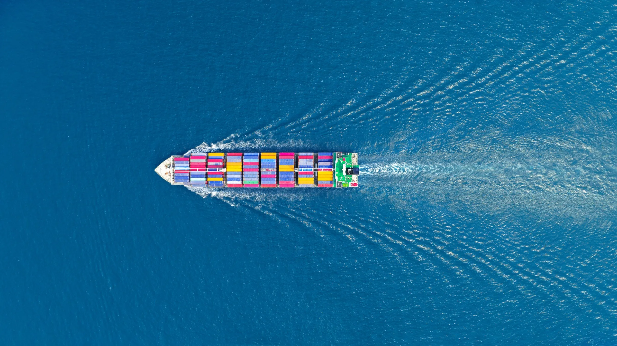 Aerial top-down view of a cargo ship fully loaded with containers, wake trailing behind